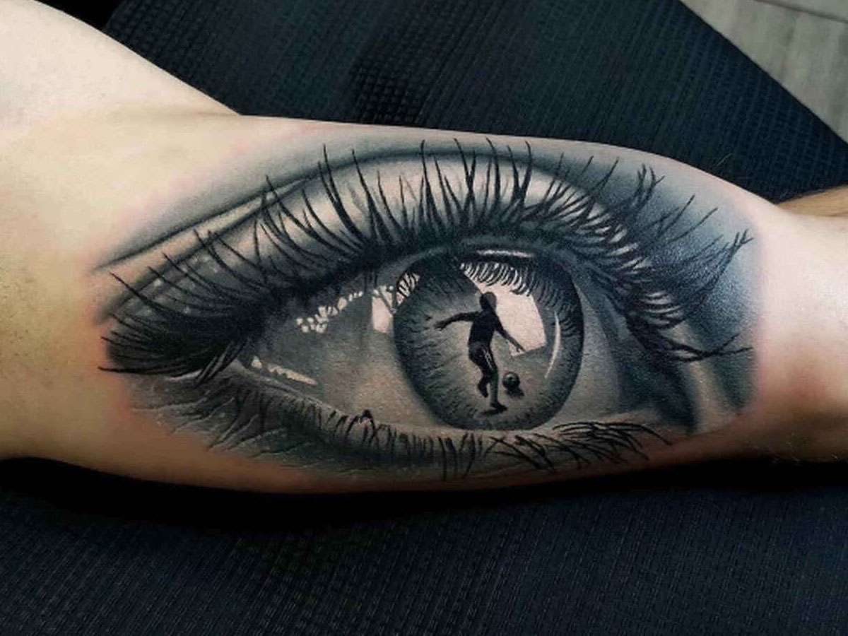 ein schwarz-graues Tattoo, das ein Auge und ein darin reflektiertes Fußball spielendes Kind zeigt